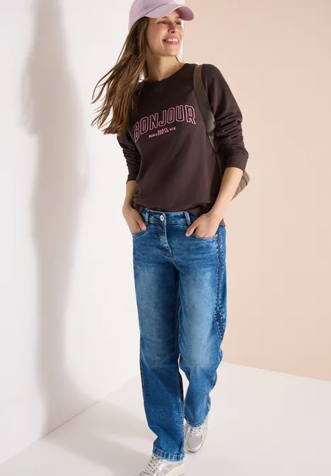 Person lehnt an Wand mit braunem Sweatshirt, blauer Jeans und Baseballcap.